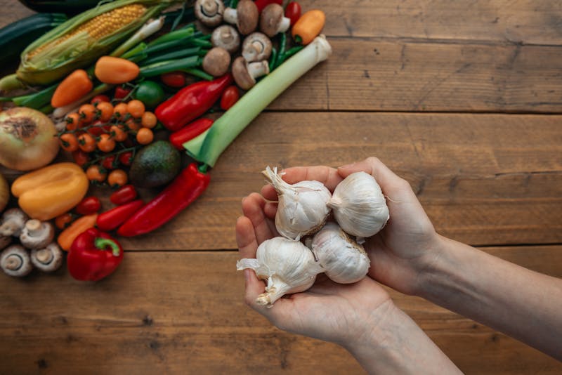 garlic bulbs held in hands, with other vegetables in the background